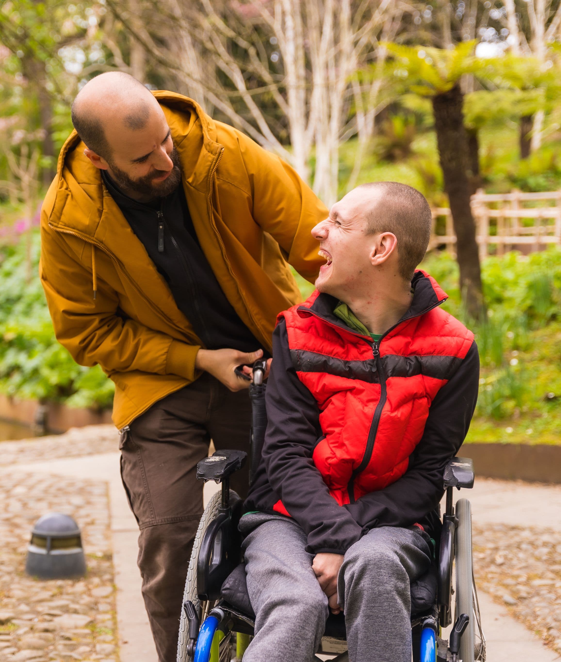 A man in a yellow jacket stands behind a man in a red vest in a wheelchair, both smiling by a pond.