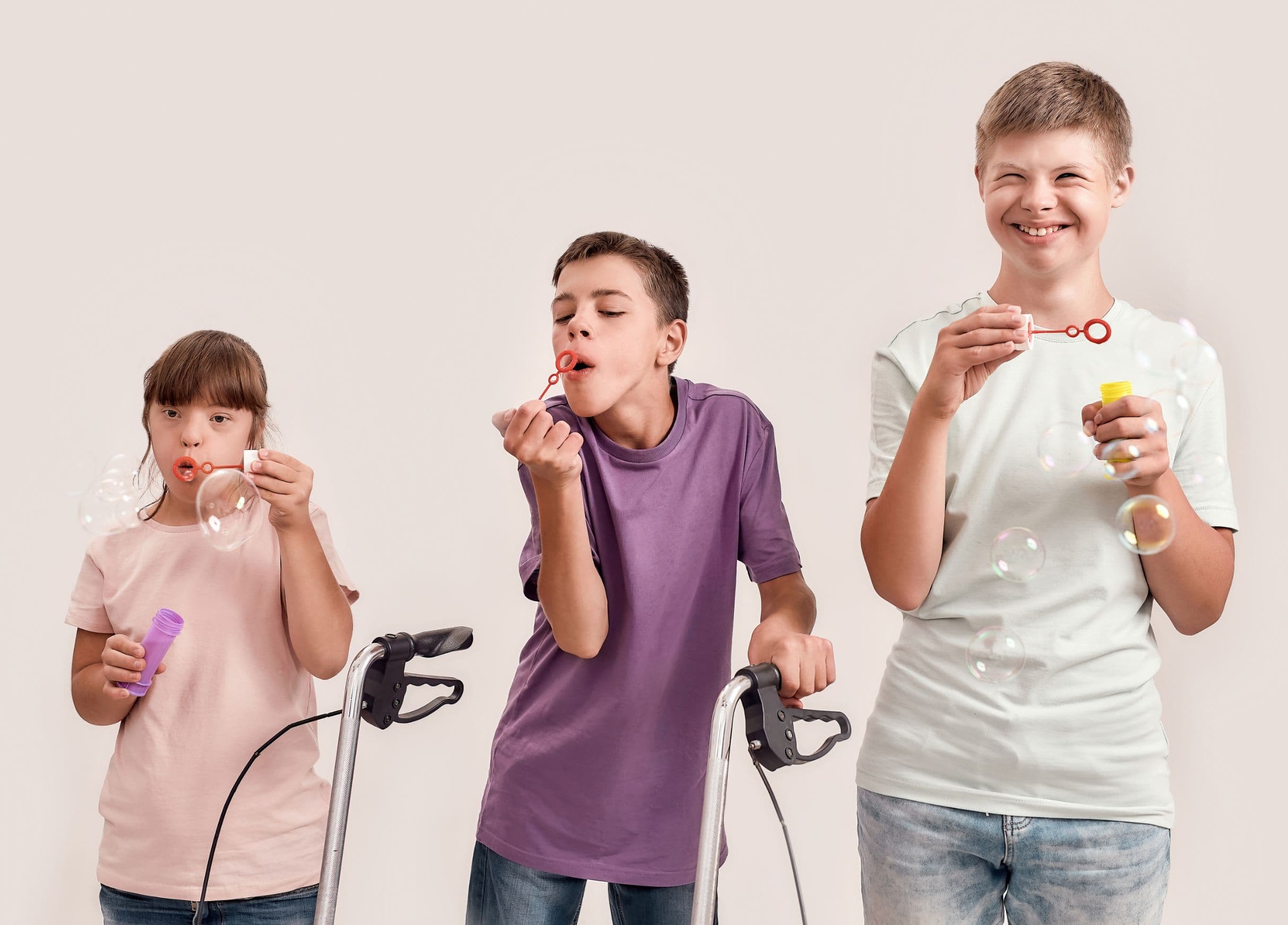 A girl and two boys using mobility walkers stand together, smiling and blowing bubbles.